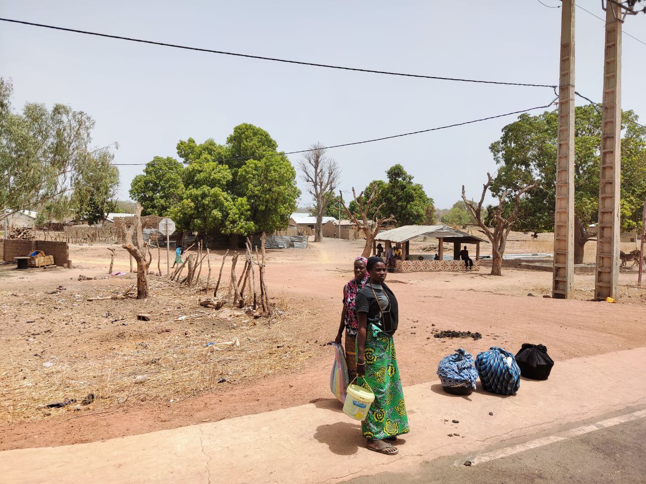 Two women with bags waiting by a roadside in the Eastern River Region, The Gambia